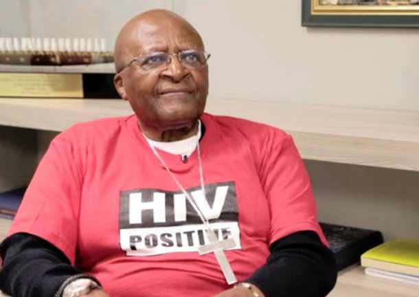 Tutu wears a red t-shirt with the words HIV positive in black and white letters. He wears a pectoral cross with necklace.