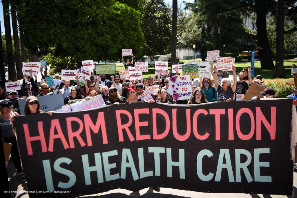 Advocates at AIDSWatch@Home march with a banner that says, "Harm Reduction Is Health Care."